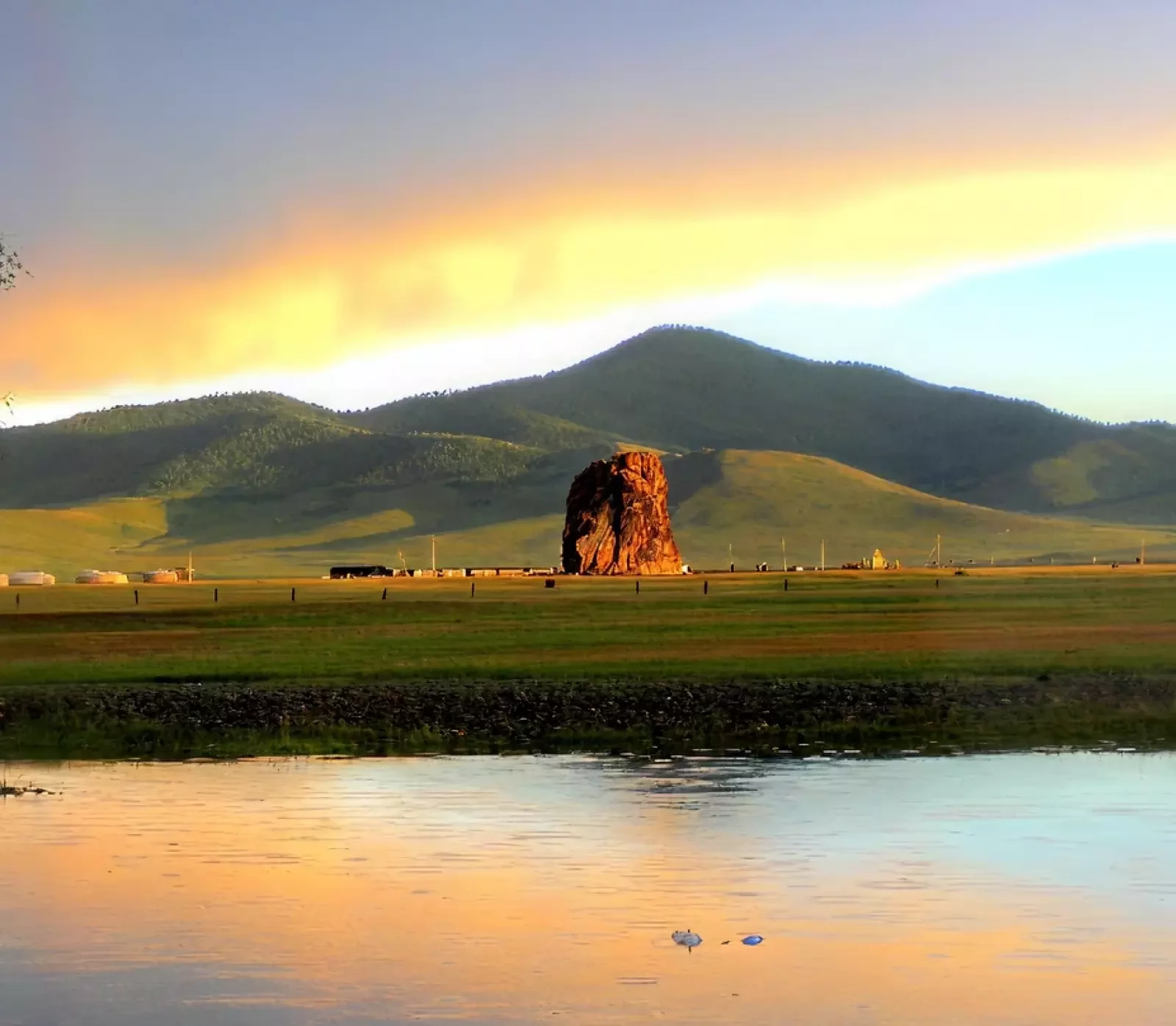 Beautiful landscape of rural Mongolia showing vast grasslands, a natural rock monument, mountain backdrop, and sunset reflections.