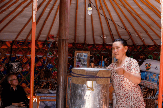 Interior of a traditional Mongolian ger with wooden roof structure and central stove, featuring a host serving tea to guests, showcasing nomadic hospitality culture