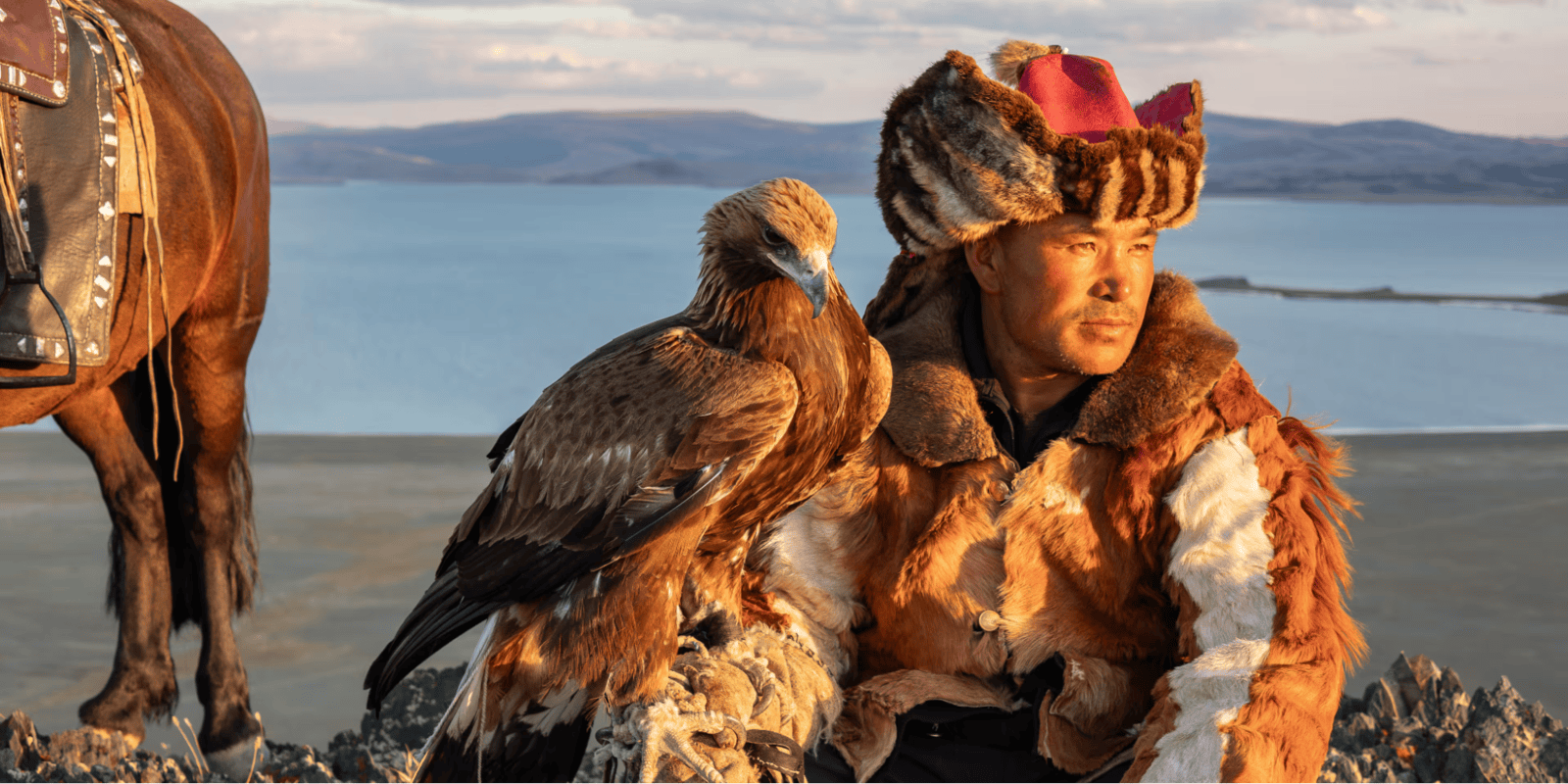 Close-up of a Kazakh eagle hunter wearing a fur hat with a golden eagle perched beside him, wings spread against a mountainous backdrop