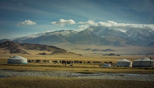 Mongolian nomadic landscape with traditional ger tents, grazing livestock, and snow-capped mountains under a clear blue sky