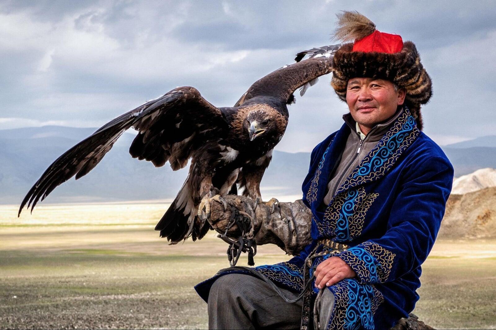 Western Mongolia eagle hunter in traditional Kazakh clothing holding a golden eagle in the Altai Mountains landscape.