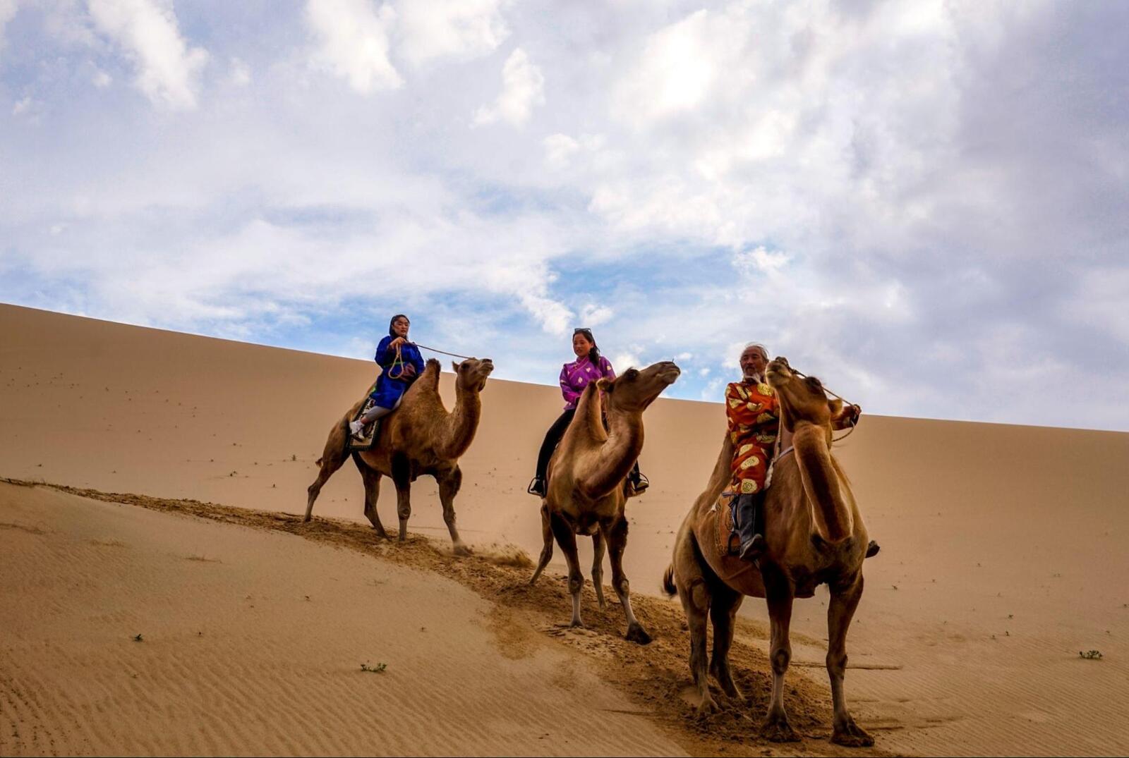 Gobi Desert camel ride with travelers crossing sand dunes under dramatic cloudy sky during Mongolia desert travel adventure.