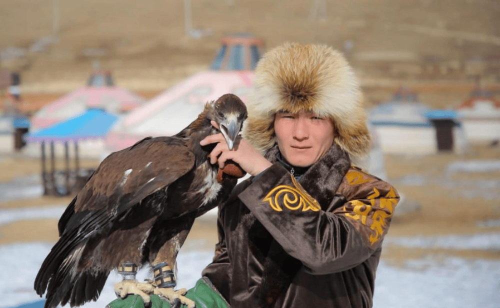 Young eagle hunter in traditional fur hat holding a golden eagle on his arm in a rural Mongolian setting