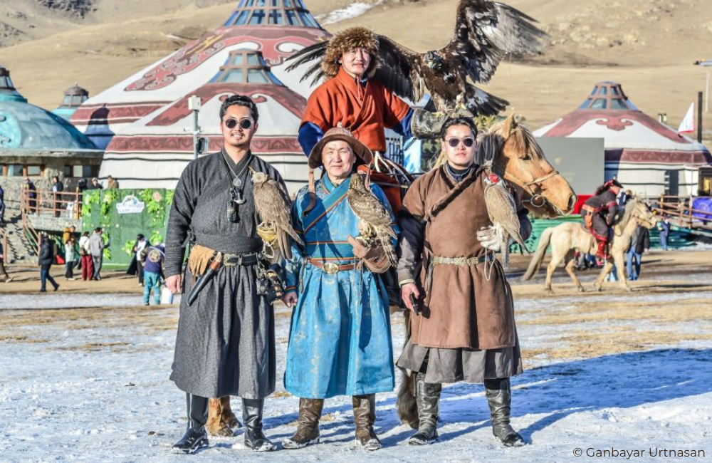 Group of Mongolian eagle hunters in traditional clothing standing in front of decorated yurts with horses and an eagle perched above during a cultural festival in the Altai region